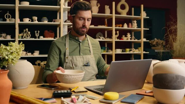 Caucasian man guy small business owner businessman crock potter artist male working measuring pottery bowl with ruler equipment making online design typing laptop computer measurement inside workshop
