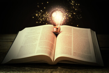 Book with glowing light bulb on wooden table and magic lights