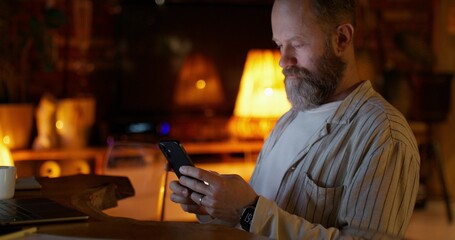 A man uses a mobile phone while working from home. He's sitting in front of an open laptop at his desk. Night work