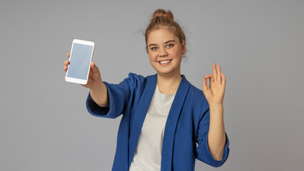 A girl in a blue jacket with a wide smile demonstrates the screen of a cell phone with the other hand shows the sign OK