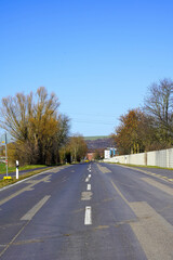 Fototapeta premium A paved road with faded markings stretches into the distance, flanked by trees and a white fence, with rolling hills in the background