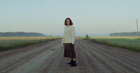 A young red-haired woman in a wet skirt stands on a gravel road among picturesque fields and looks into the camera with a confused look. 4k video, red komodo