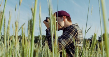 A young stylishly dressed man takes photographs with a film camera while squatting among wheat ears...