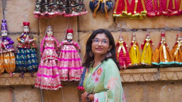 Portrait of young Indian woman looking at puppets hanging on wall of Jaisalmer local market, Rajasthan, India. Rajasthani Kathputli wearing colorful traditional Rajasthani clothes.