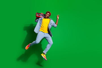 Young stylish man with a red boombox jumping joyfully against a vibrant green background in a trendy outfit