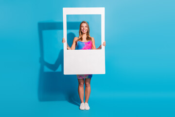 Smiling woman holding a large polaroid photo frame against a vibrant blue background wearing a...