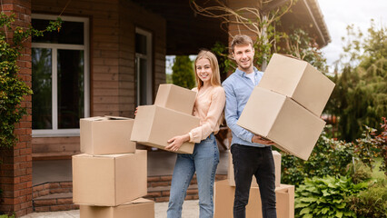 Moving to new house concept. Young guy with his wife holding cardboard boxes in front of their home
