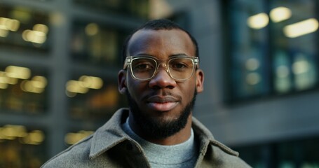 Obraz premium Stylish young African-American man smiling looking at the camera while standing near a modern building in the evening. Close-up of a man's face with glasses. Video in 4k, red komodo