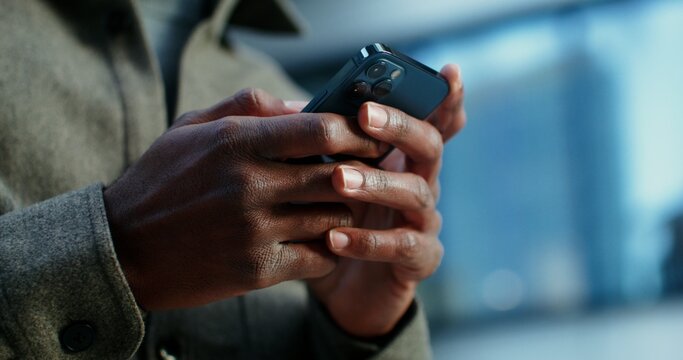 A man of African American appearance uses a mobile phone, standing near a building in which lamps shine in the windows. Close-up of male hands with a phone, no face. Video in 4k, red komodo