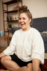 Obraz premium Crop shot of happy cheerful plus size female having fun after finishing her yoga practice at home, laughing feeling relaxed and energized, looking at camera sitting in lotus posture