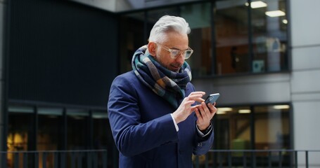 A gray-haired man of European appearance with a voluminous scarf around his neck, uses a mobile phone, standing outdoors in business center of the city among glass skyscrapers. Video in 4k, red komodo