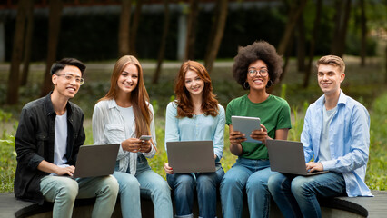 Best gadgets for college students. Multiracial teens sitting with electronic devices on bench at campus, preparing for lessons outdoors, free space