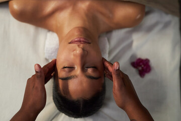 Woman receiving soothing facial massage at wellness spa while relaxing on massage table, hands of masseuse gently massaging clients forehead and cheeks