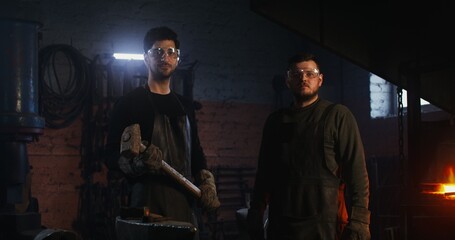 Two young blacksmiths in protective gear and goggles with working tools in their hands look at the...