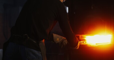 A young man in goggles and a leather apron takes a split metal billet out of the furnace with tongs...