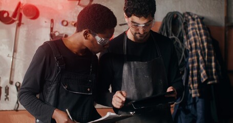 Obraz premium Two blacksmiths of different nationalities are thinking about a sketch of a future product, working together and using a tablet to search for ideas, in workshop full of tools. video in 4k, red komodo