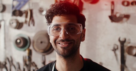 A young man in goggles smiles looking directly at the camera, against the background of a wall with...