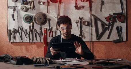 A young blacksmith in goggles and an apron draws a sketch of a future product using a tablet to search for ideas while sitting at a table in a forge. video in 4k, red komodo