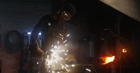 A young blacksmith of African American appearance in goggles and an apron cuts off part of a metal...