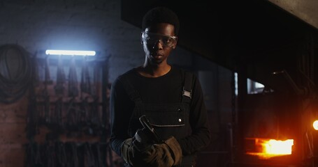 A young blacksmith of African American appearance in goggles and an apron turns on an angle grinder...