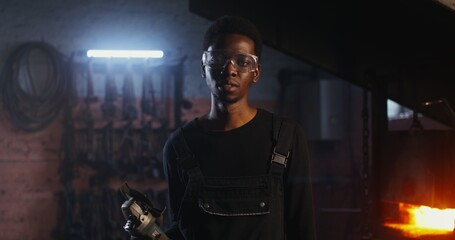 African-American man in goggles and work clothes looks at the camera with a serious look while standing in a modern forge with angle grinder in his hands. Zooming video to man's face. 4k, red komodo