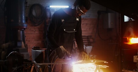 A man of African American appearance, dressed in work clothes and goggles with a concentrated look, uses a plasma cutter, cutting a sheet of iron, while working in a forge. 4k, red komodo