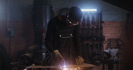 A man of African American appearance, dressed in work clothes and goggles with a concentrated look, uses a plasma cutter, cutting a sheet of iron, while working in a forge. 4k, red komodo