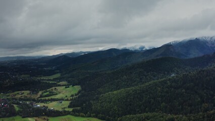Naklejka premium Vertical panning from a drone, a view of mountain valley with city in a lowland at foot of gently sloping mountains overgrown with a spruce forest. Rocky peaks are covered with clouds, on a summer day
