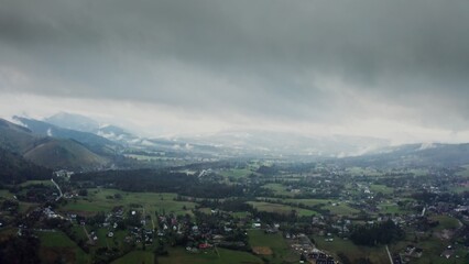 Fototapeta premium Vertical panning from a drone, a view of mountain valley with city in a lowland at foot of gently sloping mountains overgrown with a spruce forest, on a cloudy day at dusk