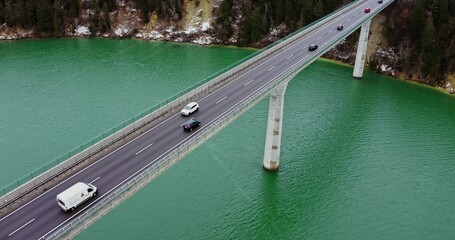Top view of a road bridge with a two-lane asphalt road with cars passing on it. drone video footage of a long bridge across an emerald green river to hills covered in snowy forest.