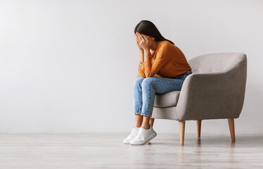 Young Asian woman burying her face in hands and crying, feeling depressed, sitting in armchair...