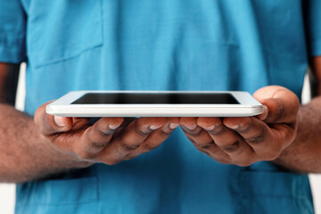 A millennial African American man doctor stands isolated against a plain background, holding a tablet to enhance patient care and engage with medical information efficiently.