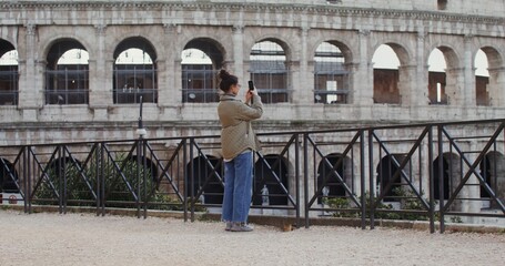 A young warmly dressed woman of European appearance takes photographs of the sights of Rome on a mobile phone, standing on an observation deck near the Coliseum. Video in 4k, red komodo.