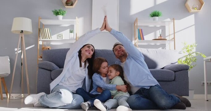 Happy young smiling parents mother, father with children sitting on floor at home making house roof by hands above. Insurance, care,love and family protection concept. 4k video. Slow motion video.