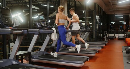 Young man and woman of European appearance, sporty physique, dressed in stylish sportswear, run on a treadmill, working out in a modern gym together