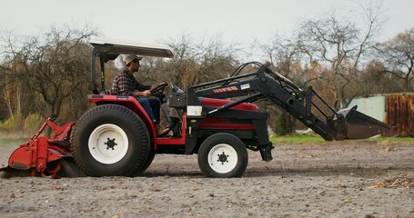 Obraz premium A young male tractor driver drives a modern multipurpose agricultural machine plowing a field on an autumn day. Side videography