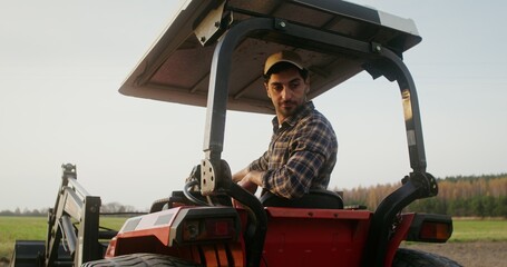 A young European farmer operates a farm tractor, while sitting in an open cab with canopy and...