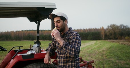 Young black-haired male farmer with a beard eating a sandwich while standing near a modern...