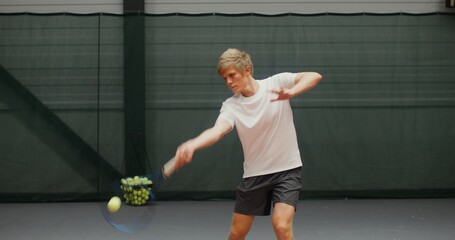 Obraz premium A young man of european appearance with a concentrated look hits a tennis ball, with a racket while playing with his partner in an indoor tennis court
