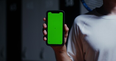 A young man with a racket in his hand holds a phone with a green screen, standing in the locker room of the sports hall. Close-up phone, man's face is not visible, copy space
