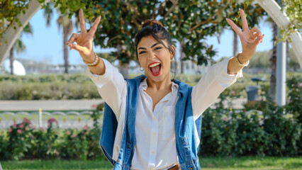 A young brunette businesswoman in a tree-filled park on a spring day, making a victory sign with...