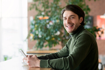 Obraz premium Young handsome guy using mobile phone at cafe, smiling at camera, copy space