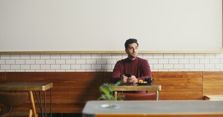 A young dark-haired man with a slight unshaven sits at a small table in a cafe with a mug of coffee...