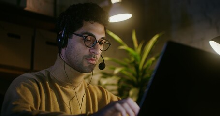 Young dark-haired man with glasses, with stubble on his cheeks, talking on a microphone in headphones while sitting in front of an open laptop under the light of lamps