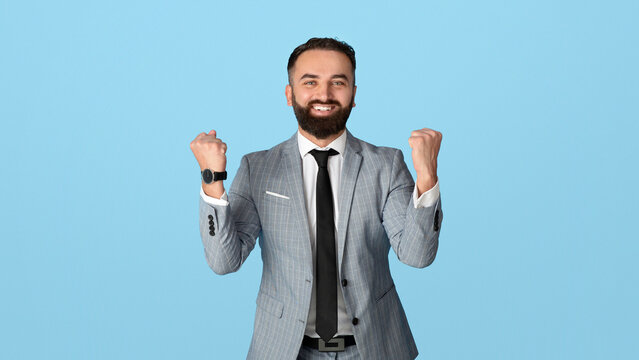 Handsome young businessman feeling excited and gesturing YES on blue studio background. Attractive millennial office employee celebrating his success, feeling triumphant