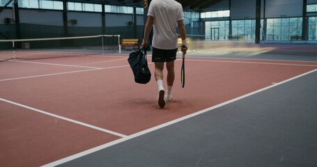 Obraz premium A young man in a sports uniform walks into an indoor tennis court with a racket and a bag of things, going to play tennis, video filming from the back
