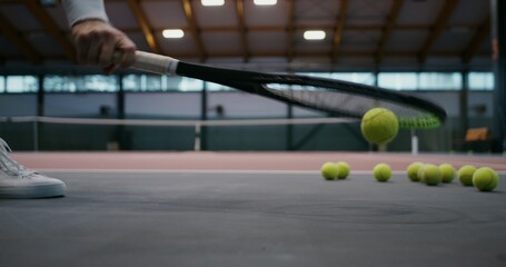 A lot of tennis balls lie on the rubberized floor of an indoor tennis court, a man picks up balls...