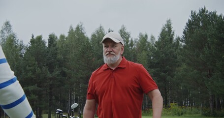 An elderly gray-haired man with a beard in a baseball cap, dressed in stylish sportswear, talks to another man while standing on a golf course. Outdoor video shooting in daylight