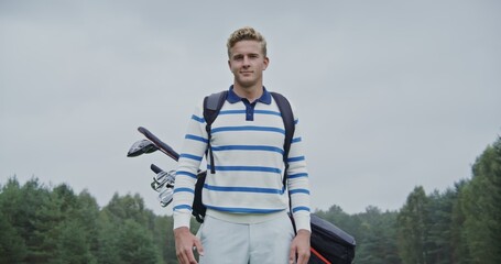 A young handsome blond man of European appearance, dressed in sportswear with golf equipment in a bag behind his back, stands on the golf course and looks at the camera smiling
