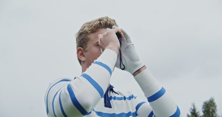 A young handsome man in light-colored clothes and a white golf glove peers through binoculars in...